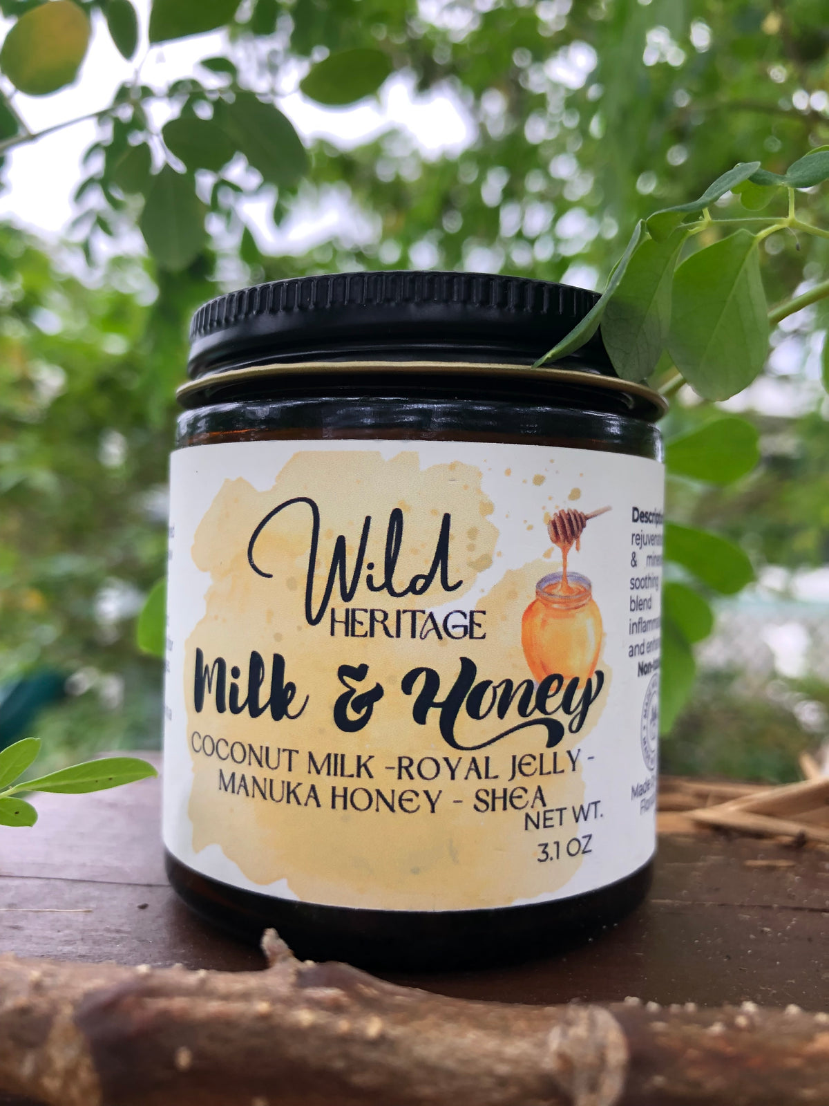 A jar of 'Milk & Honey' coconut milk and royal jelly face balm, with a black lid and white label, placed on a wooden surface with flowers in the background.